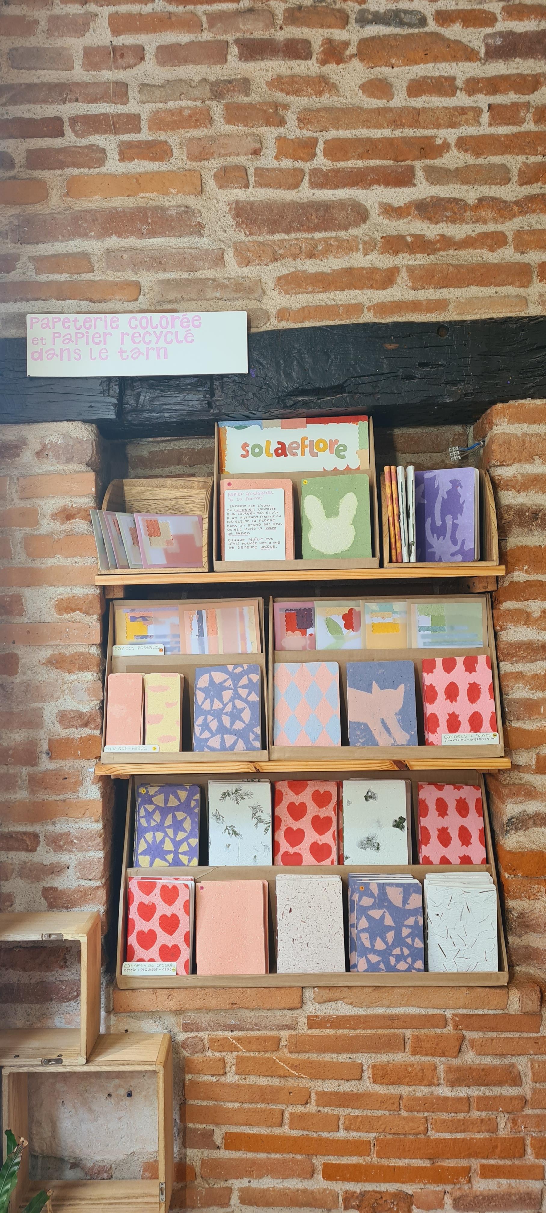 Étagères en intérieur sur 3 niveaux avec des cartes postales, des carnets colorés et des marque-pages. Tout en haut une pancarte avec écrit "solaeflore" en lettres découpées en papier recyclé. Étagères en intérieur sur 3 niveaux avec des cartes postales, des carnets colorés et des marque-pages. Tout en haut une pancarte avec écrit "solaeflore" en lettres découpées en papier recyclé.