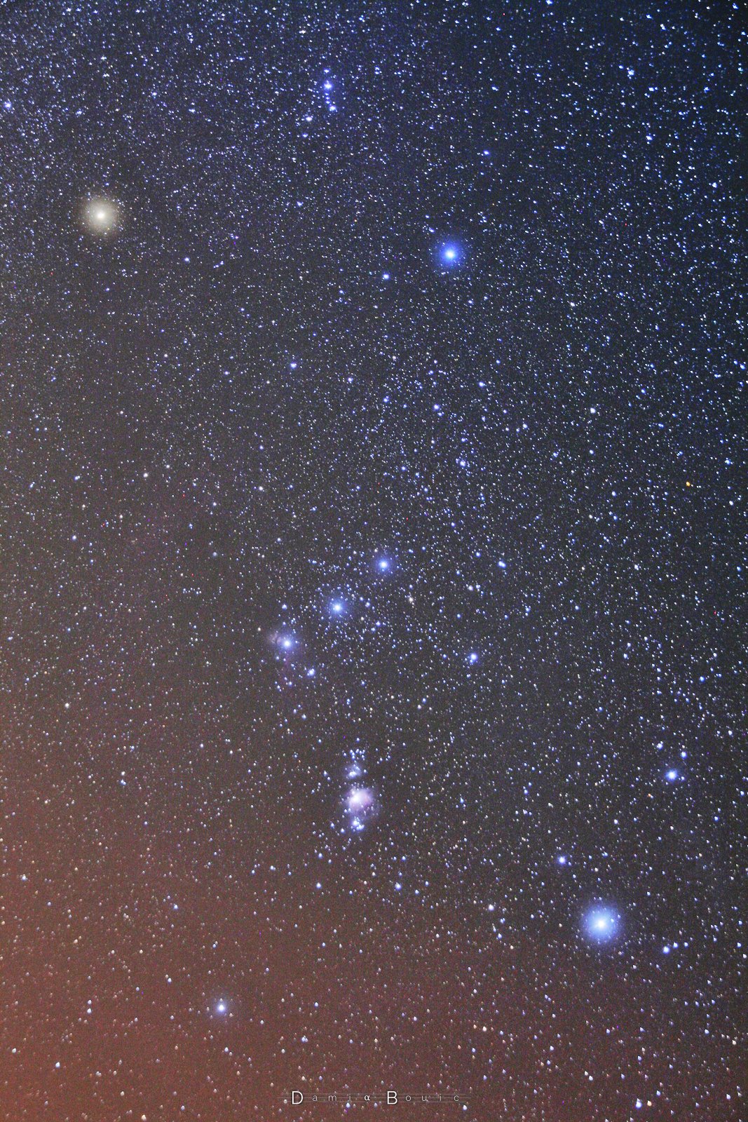 Image montrant plein plein d'étoiles, dont 7 visibles sous forme de points lumineux diffus. Ceux-ci sont arrangés de façon à former deux trapèzes collés par leur base la plus courte. Au centre, à cette zone collée, trois étoiles, de luminosité égale, dont celle de gauche est flanquée d'une toute petite touche de pinceau doux, légèrement rouge.
Dans le trapèze du bas, au centre et dans le sens vertical, un groupement d'étoiles, et au milieu, un forme floue : la nébuleuse d'orion.