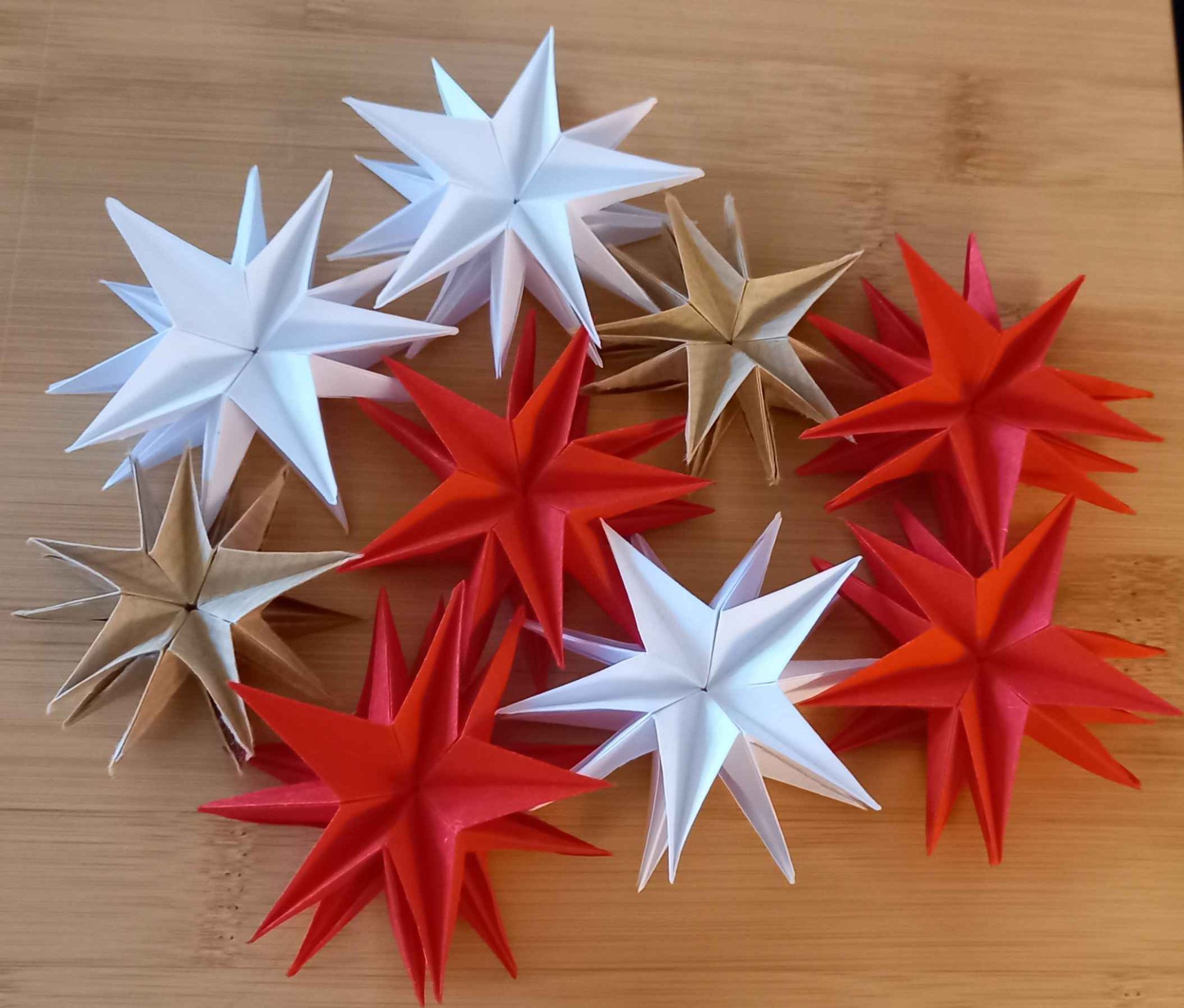 9 étoiles en origami de tailles et de couleurs différentes (en papier blanc, rouge, craft), posées sur une table en bois. 9 étoiles en origami de tailles et de couleurs différentes (en papier blanc, rouge, craft), posées sur une table en bois.