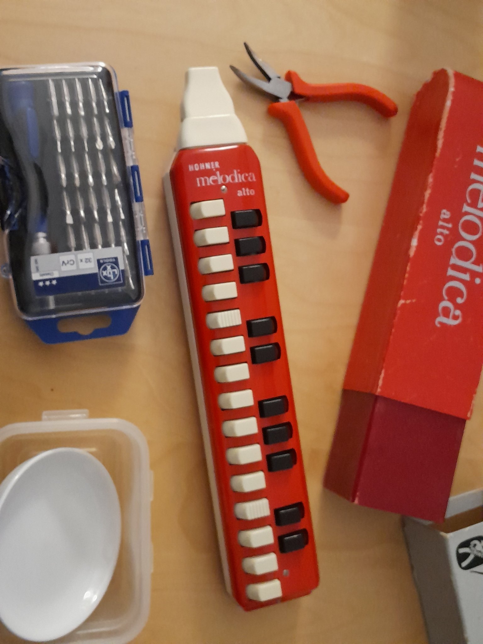 un mélodica alto de la marque Hohner rouge avec sa boite d'origine, à coté d'une boite de tournevis de petite taille, une paire de pinces, deux tupperwares et une petite assiette. un mélodica alto de la marque Hohner rouge avec sa boite d'origine, à coté d'une boite de tournevis de petite taille, une paire de pinces, deux tupperwares et une petite assiette.