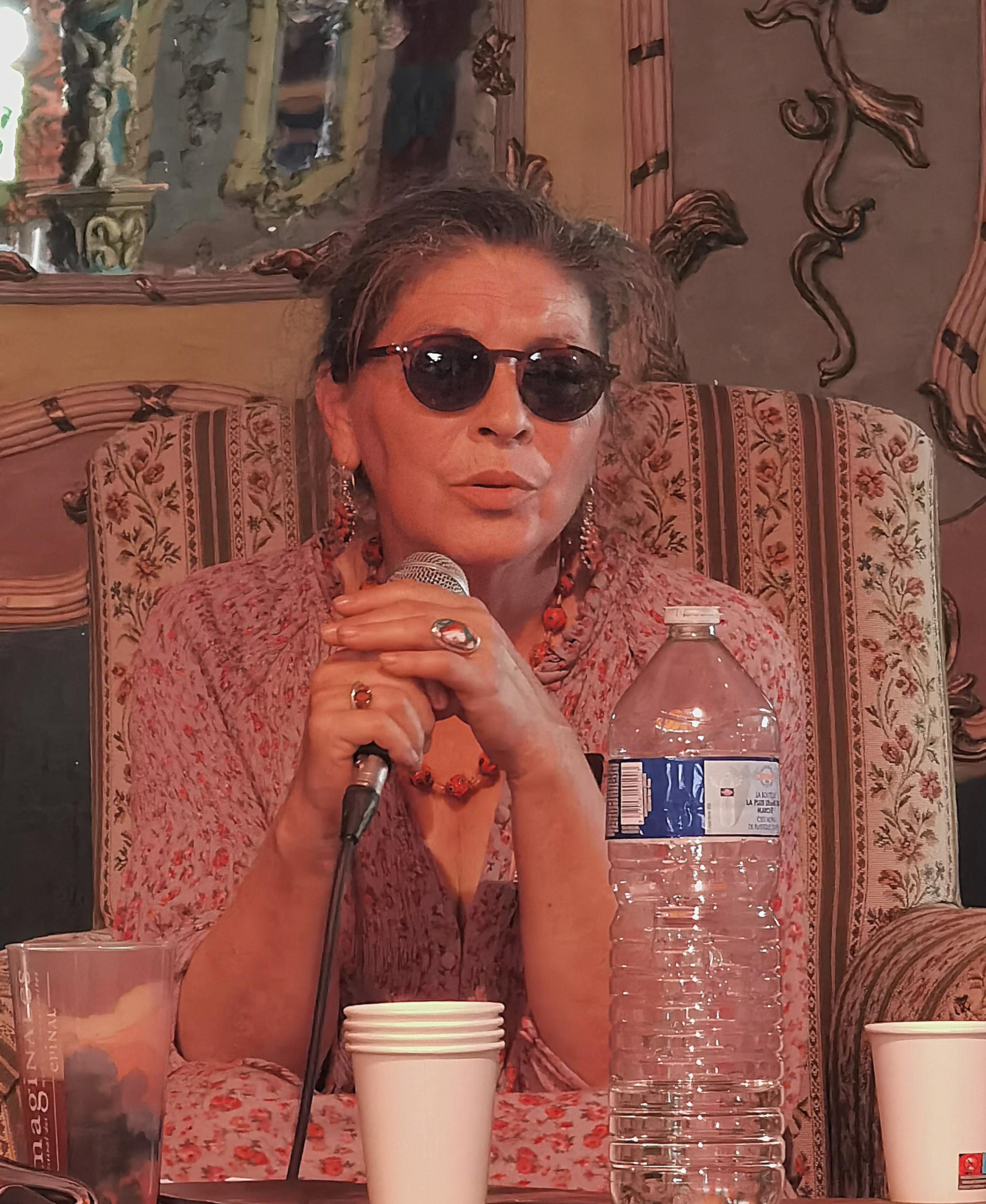 conférence aux Imaginales 2026. Assise derrière une table sous le chapiteau, je tiens un micro entre mes mains tandis que je m’adresse au public. Je porte des lunettes de soleil noires, une robe fleurie rose et des bijoux orange . Devant moi s’alignent une grande bouteille d’eau et plusieurs gobelets, indispensables pour affronter les 40 °C qui règnent sous la tente. Le décor aux accents baroques et les fauteuils à motifs donnent à la scène une atmosphère chaleureuse et un peu hors du temps. conférence aux Imaginales 2026. Assise derrière une table sous le chapiteau, je tiens un micro entre mes mains tandis que je m’adresse au public. Je porte des lunettes de soleil noires, une robe fleurie rose et des bijoux orange . Devant moi s’alignent une grande bouteille d’eau et plusieurs gobelets, indispensables pour affronter les 40 °C qui règnent sous la tente. Le décor aux accents baroques et les fauteuils à motifs donnent à la scène une atmosphère chaleureuse et un peu hors du temps.