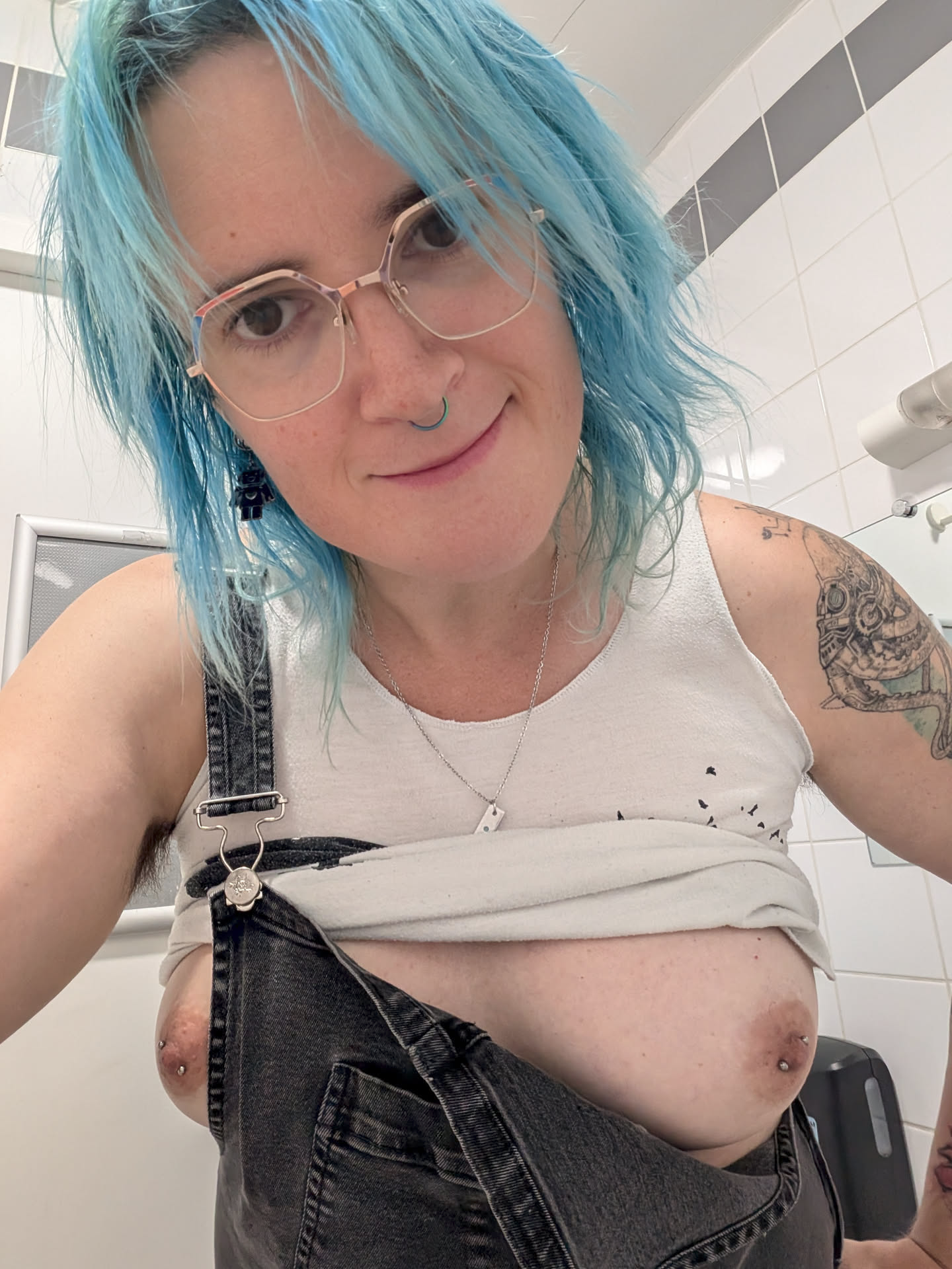 A picture of Miranda, a white fem person with blue hair, standing in a public toilet. They're wearing a black dungaree over a white top, but one strap of the dungaree is undone and the top is raised above their boobs. They're smiling at the viewer. A picture of Miranda, a white fem person with blue hair, standing in a public toilet. They're wearing a black dungaree over a white top, but one strap of the dungaree is undone and the top is raised above their boobs. They're smiling at the viewer.