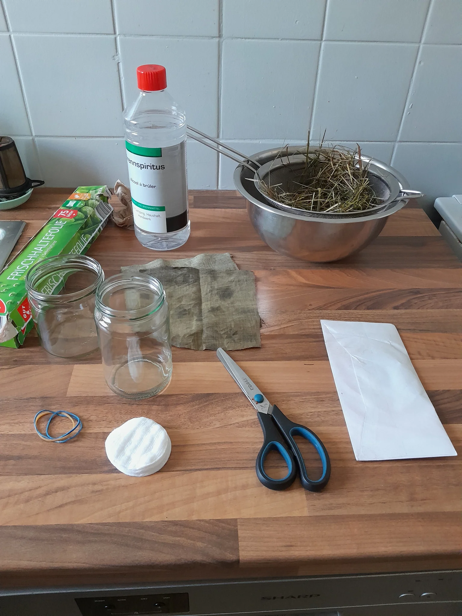 Sur un plan de travail, une paire de ciseaux, une enveloppe, des boceaux en verre, des carrés de tissu, du film alimentaire, de l'alcool à brûler et de la paille humide dans un saladier. Sur un plan de travail, une paire de ciseaux, une enveloppe, des boceaux en verre, des carrés de tissu, du film alimentaire, de l'alcool à brûler et de la paille humide dans un saladier.