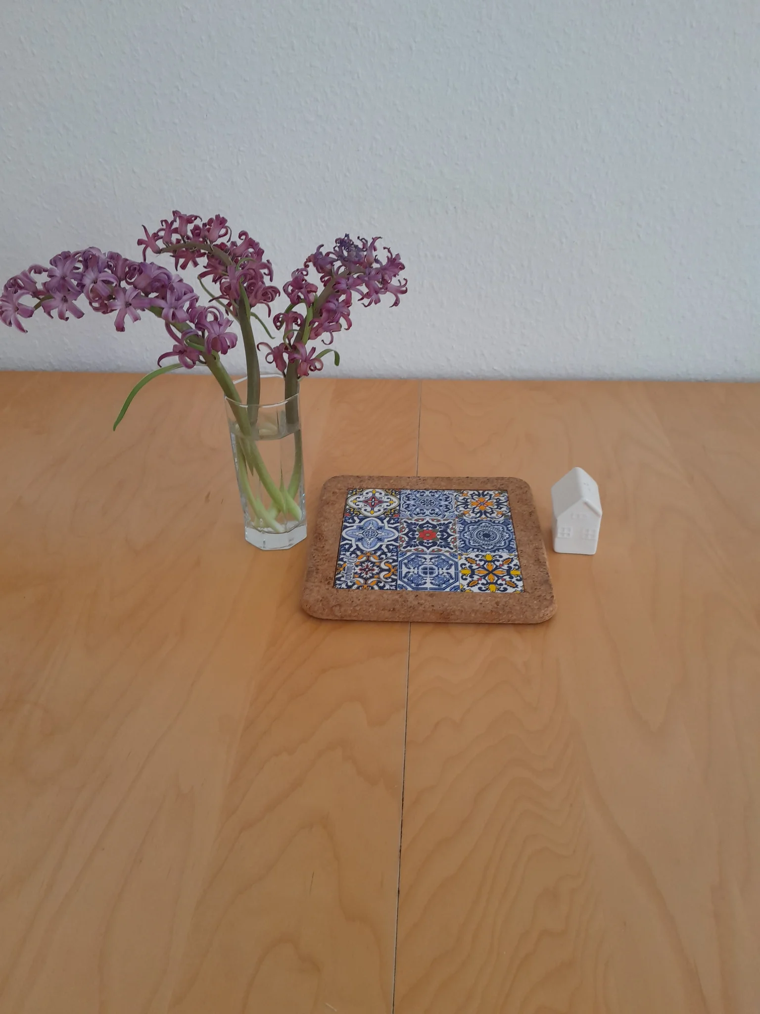 Sur une table propre et vide. Un joli verre octogonal avec trois tiges de Jacynthes coupées, un dessous de plat avec une céramique de carreaux de style facades portugaises et une salière en céramique blanche en forme de petite maison. Sur une table propre et vide. Un joli verre octogonal avec trois tiges de Jacynthes coupées, un dessous de plat avec une céramique de carreaux de style facades portugaises et une salière en céramique blanche en forme de petite maison.
